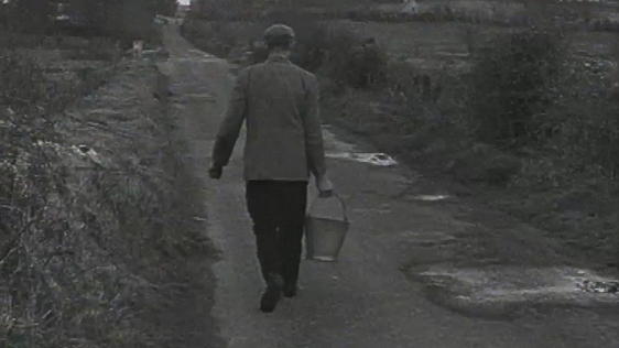 A farmer carries a bucket of water along a country road.