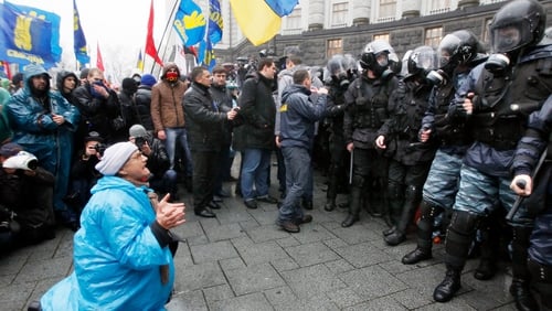 An elderly woman kneels before a line of riot police