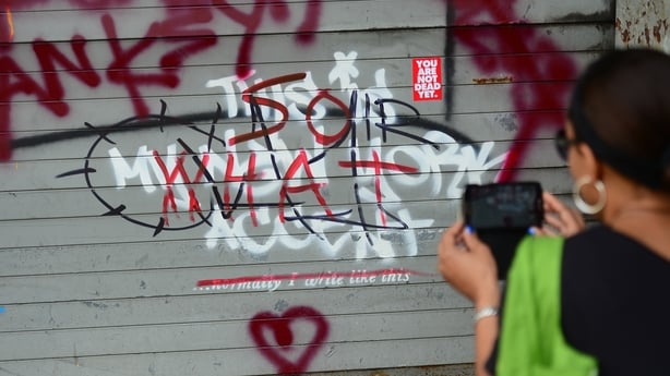 A woman takes a picture of a defaced street art graffiti by elusive artist Banksy
