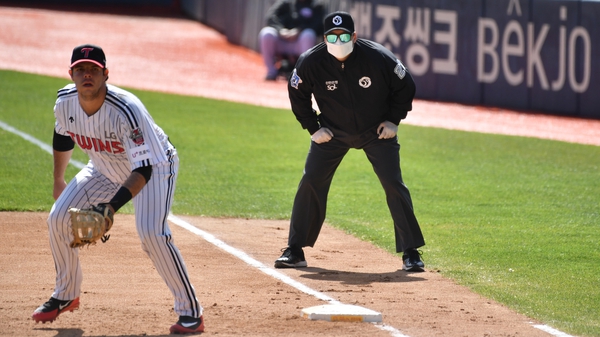 A referee (R) wearing a face mask looks on at first base