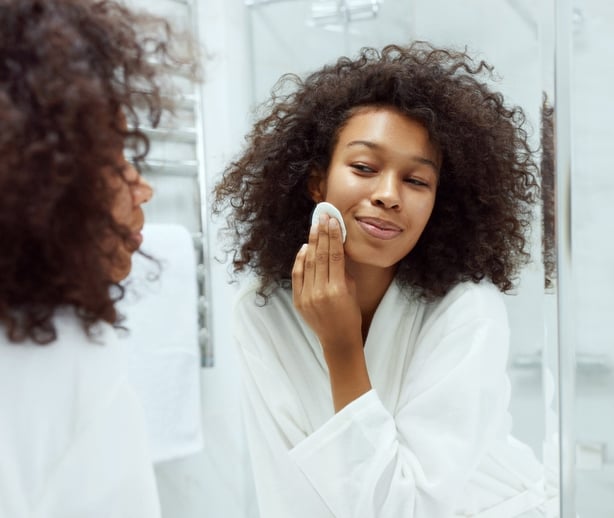 Face skin care. Girl removing makeup with cosmetic cotton pad at bathroom. Closeup portrait of beautiful smiling african woman with afro hair cleaning facial skin with white pad