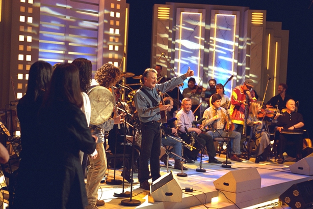 Irish musician Dónal Lunny and other guest musicians, on the set of RTÉ Television's 'The Late Late Show' in April 1996. He is standing in the centre of a large group of musicians, in right profile facing out towards the audience. Other musicians on the stage include Christy Moore, Liam O'Flynn, Paddy Glacken and Liam Ó Maonlaí. Photo by Thomas Holton