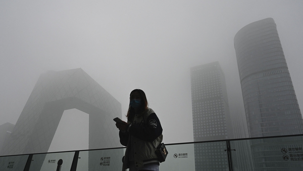 A woman uses her phone along a glass wall with the China Central Television (CCTV) headquarters in the background on a polluted day in Beijing