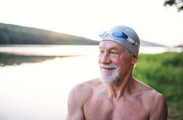 A mature man next to a lake wearing a swimming cap and goggles