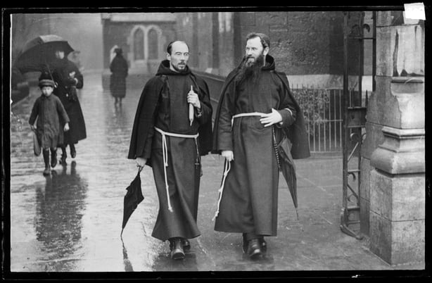 Father Albert Bibby (left) and Father Dominic O'Connor both wear priests' robes, circa 1920. In the background is a church, most likely the Capuchin Friary and Church Street Church in Dublin .