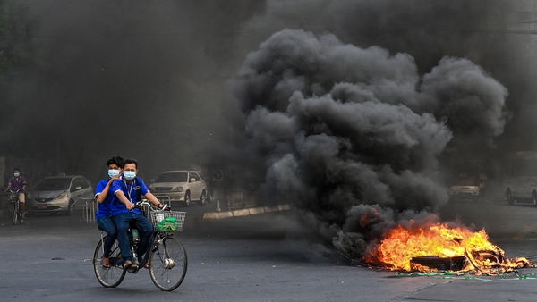 This picture taken on 3 April, 2021, shows people cycling past a burning makeshift barricade, erected by protesters demonstrating against the military coup