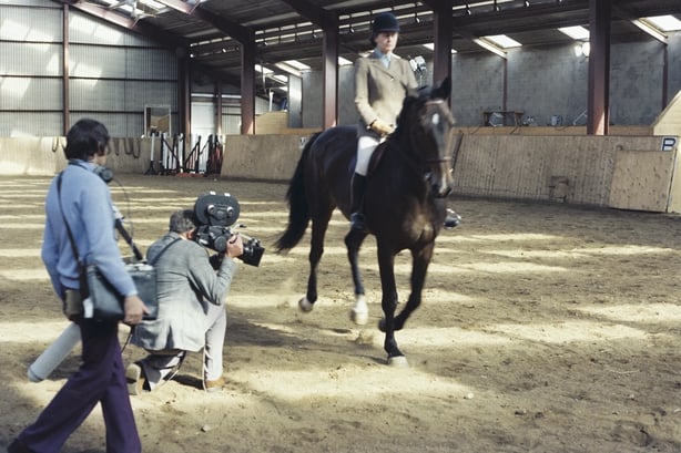 A colour photo of Iris Kellett in riding gear riding a horse in an area, filmed by a TV camera