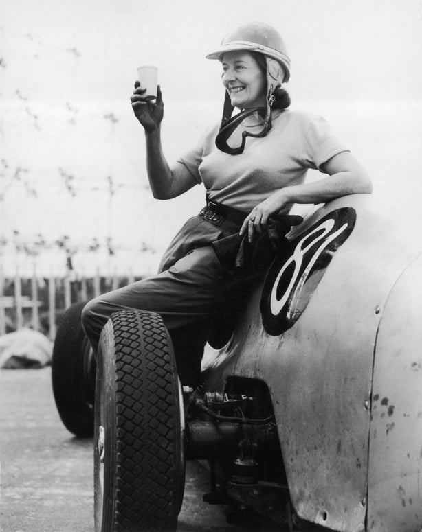 A smiling Taylour raises a glass while wearing a racing helmet