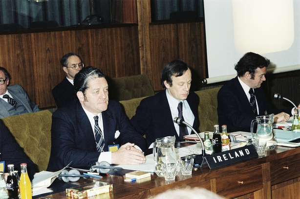 Colour photo of men sitting at a desk behind microphones