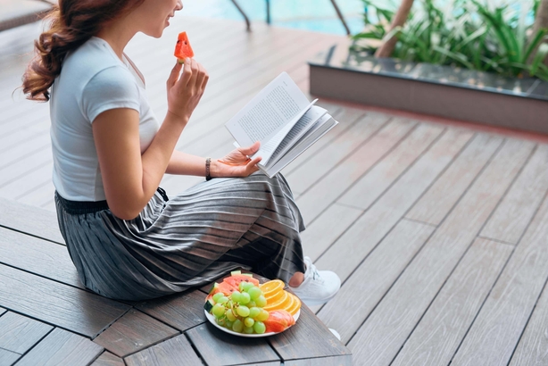 Woman sitting by a pool, eating a platter of fruit while reading a book