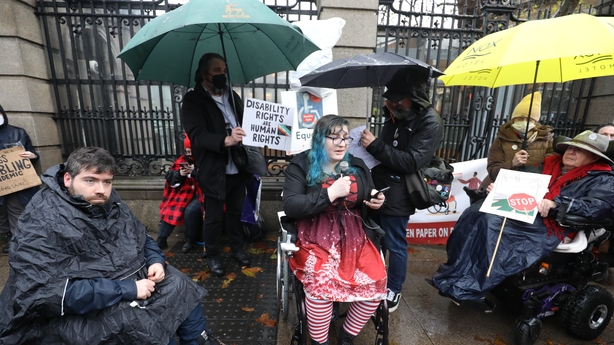 Photo of protester Protesters in the rain. One wheelchair user is holding a microphone. Behind her a protester is holding a placard that says 'Disability Rights and Human Rights'.