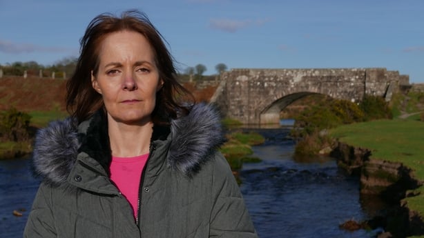 A colour photo of a white woman wearing a parka in front of a bridge