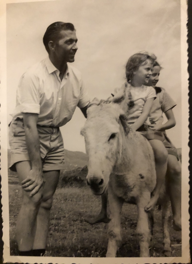 A black and white photo of a smiling man with two small children sitting on a donkey