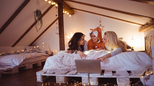 Women in pjs laying on the bed in the bedroom decorated with string lights, watching movies online streaming