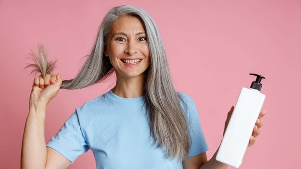 Woman with long grey hair holding a bottle of hair product, standing against a pink background, looking happy and confident