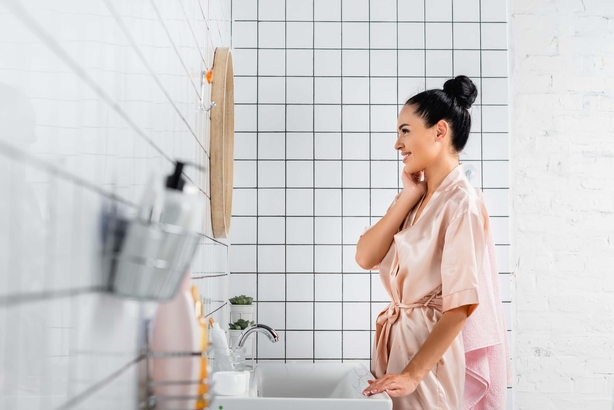 Young woman smiling while looking at mirror near toiletries on blurred foreground