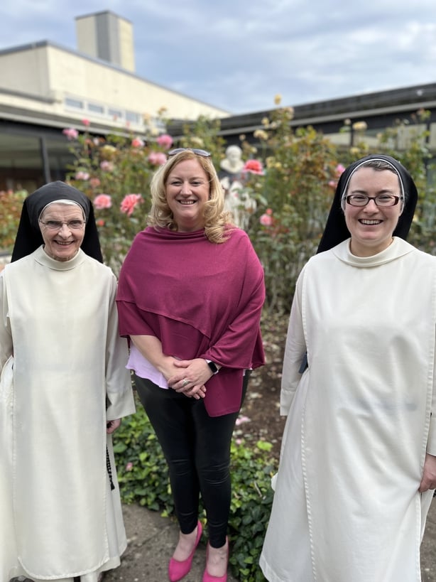 Color photo of Dearbail MacDonald, a blonde in a pink top, standing between two nuns dressed in white.