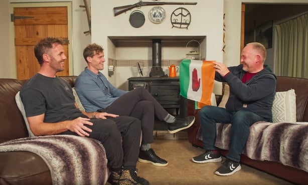 Andrew Trimble and Barry Murphy, two white young men, sit on a couch on the left. Facing them another white man, Mark Thompson, holds up an Irish tricolour with a red hand in the white panel