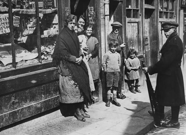 A group of women stand by a shop doorway in 1920s clothes while a soldier looks on