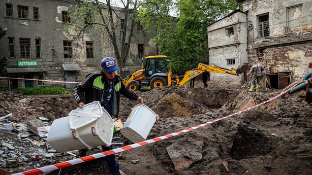 An employee of a medical facility carries things out of a surviving building against the backdrop of a sinkhole from a Russian missile hit.
