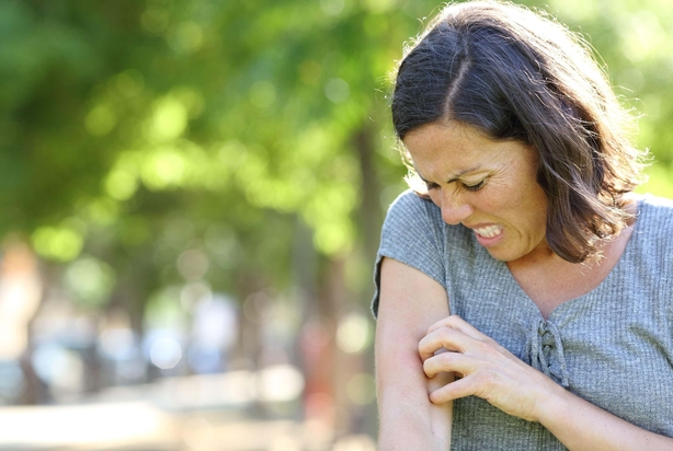 Adult woman with itchy skin scratches arm standing in the park at summer