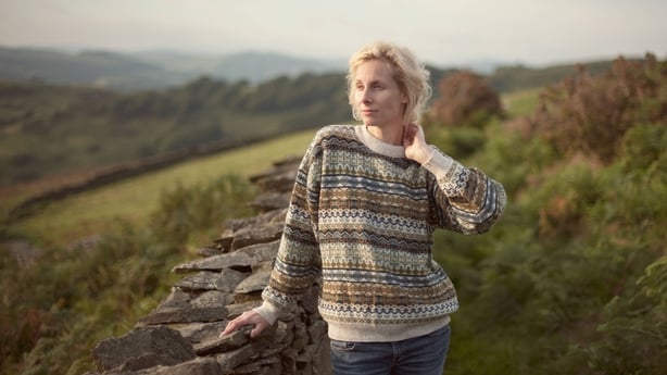 A young woman in a jumper and jeans stands by a stone wall in the countryside with green fields