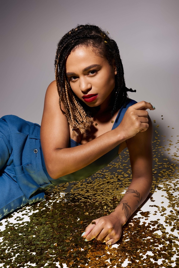 A chic young woman with braided hair and vibrant red lips showcases her unique style against a shimmering backdrop.