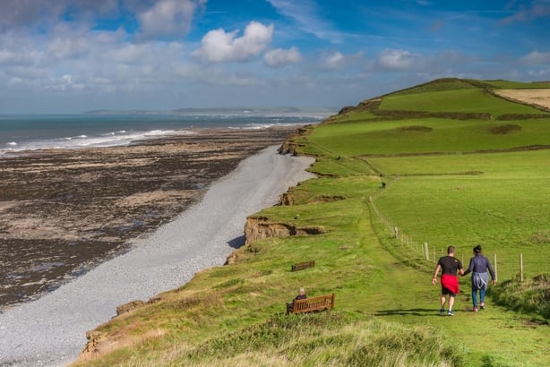 A couple walk along the South West Coast Path, looking towards Westward Ho!, as the sunshine lights up the footpath.